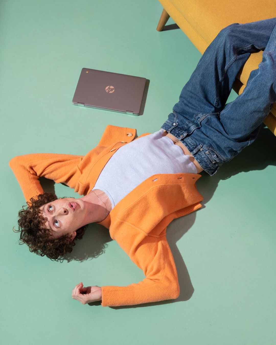 Young adult male lies on the green floor next to a laptop wearing jeans, a white t-shirt, and an orange cardigan with a yellow chair nearby.