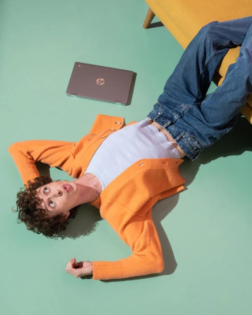 Young adult male lies on the green floor next to a laptop wearing jeans, a white t-shirt, and an orange cardigan with a yellow chair nearby.
