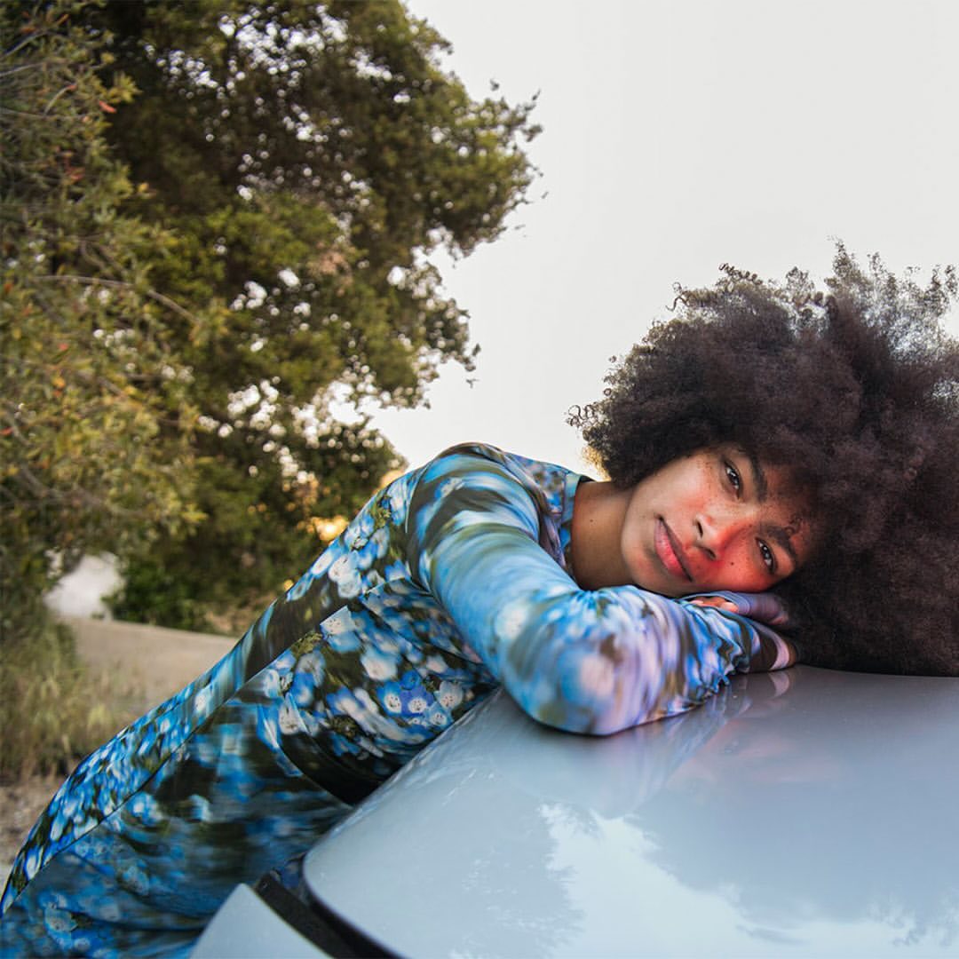 A woman with dark curly hair rests her head on a silver car hood outdoors, wearing a long-sleeved blue and white patterned top near trees.