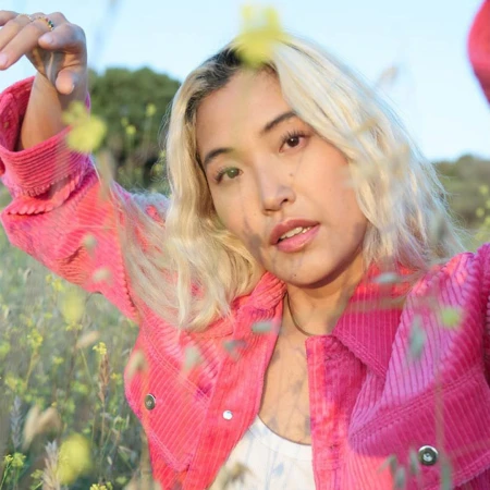 Close-up portrait of a woman with blonde hair wearing a bright pink corduroy jacket outdoors among wildflowers.