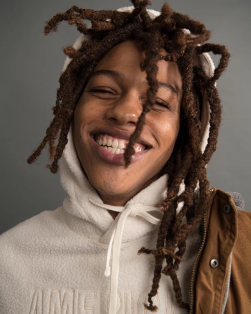 Close-up studio shot of a smiling adult male with dreadlocks wearing a hooded sweatshirt and jacket, set against a neutral grey background.