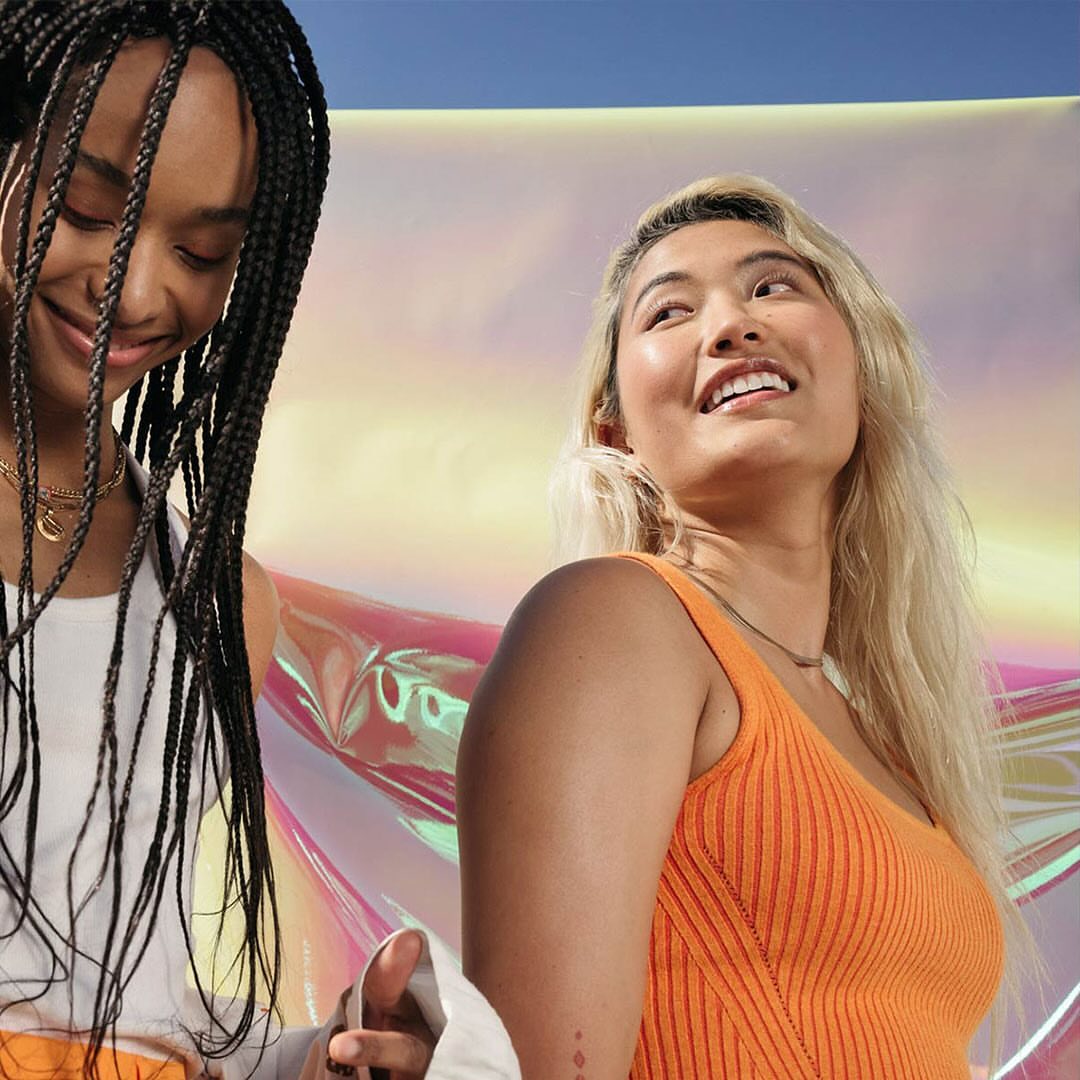 Two smiling women, one with blonde hair and the other with black braids, stand indoors against an iridescent background in a medium shot.