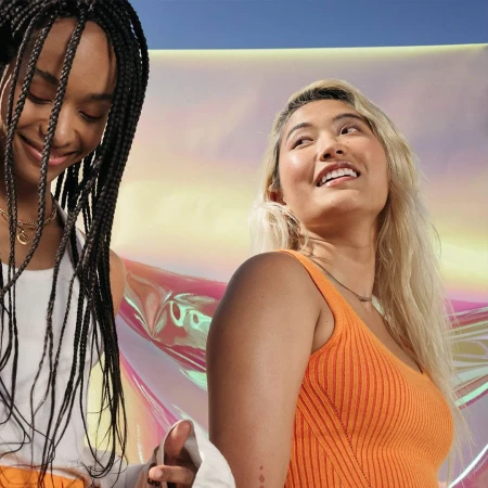 Two smiling women, one with blonde hair and the other with black braids, stand indoors against an iridescent background in a medium shot.