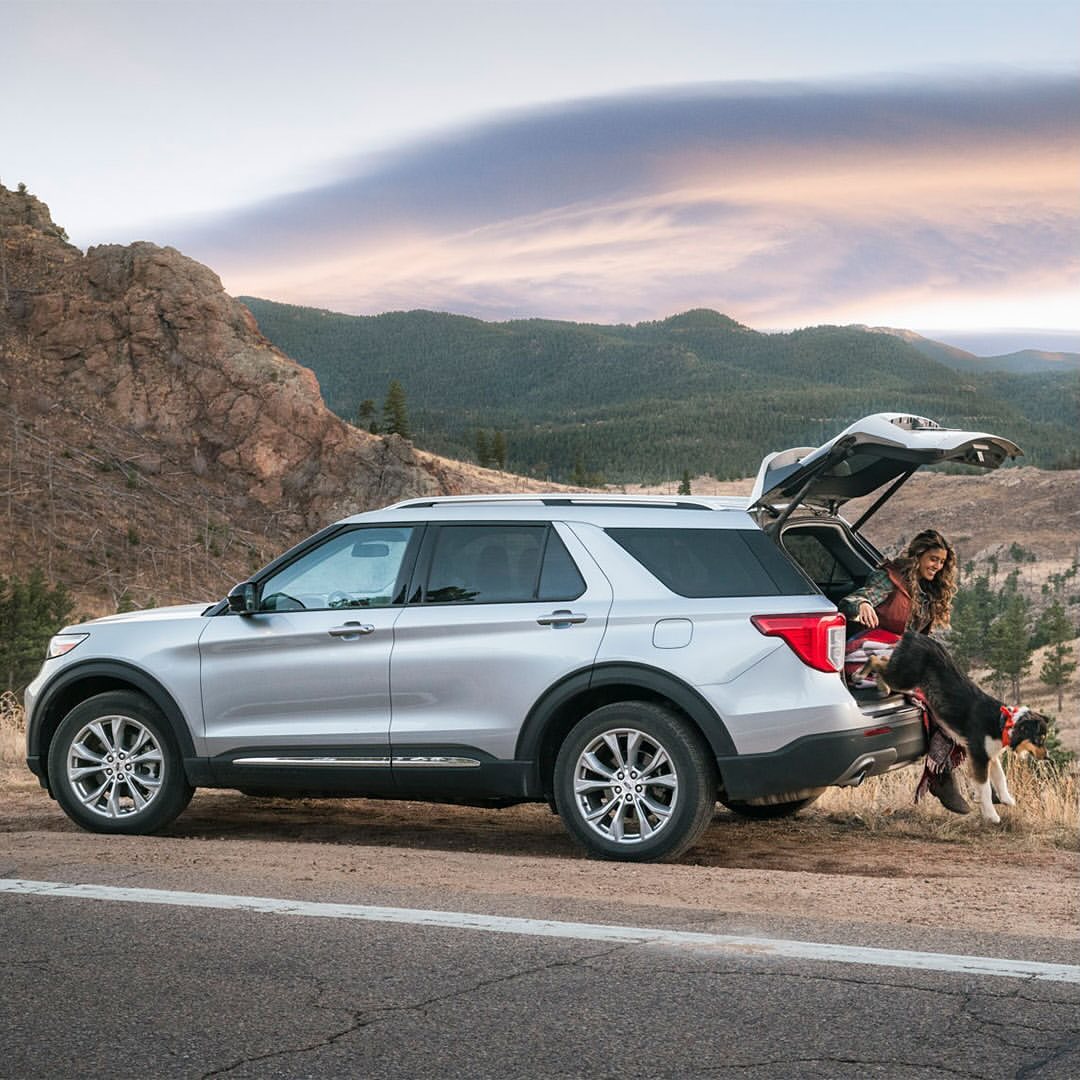 Silver Ford Explorer SUV parked on the roadside with a woman and dog at the open trunk, backed by scenic mountain views and dusk sky.