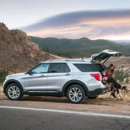 Silver Ford Explorer SUV parked on the roadside with a woman and dog at the open trunk, backed by scenic mountain views and dusk sky.