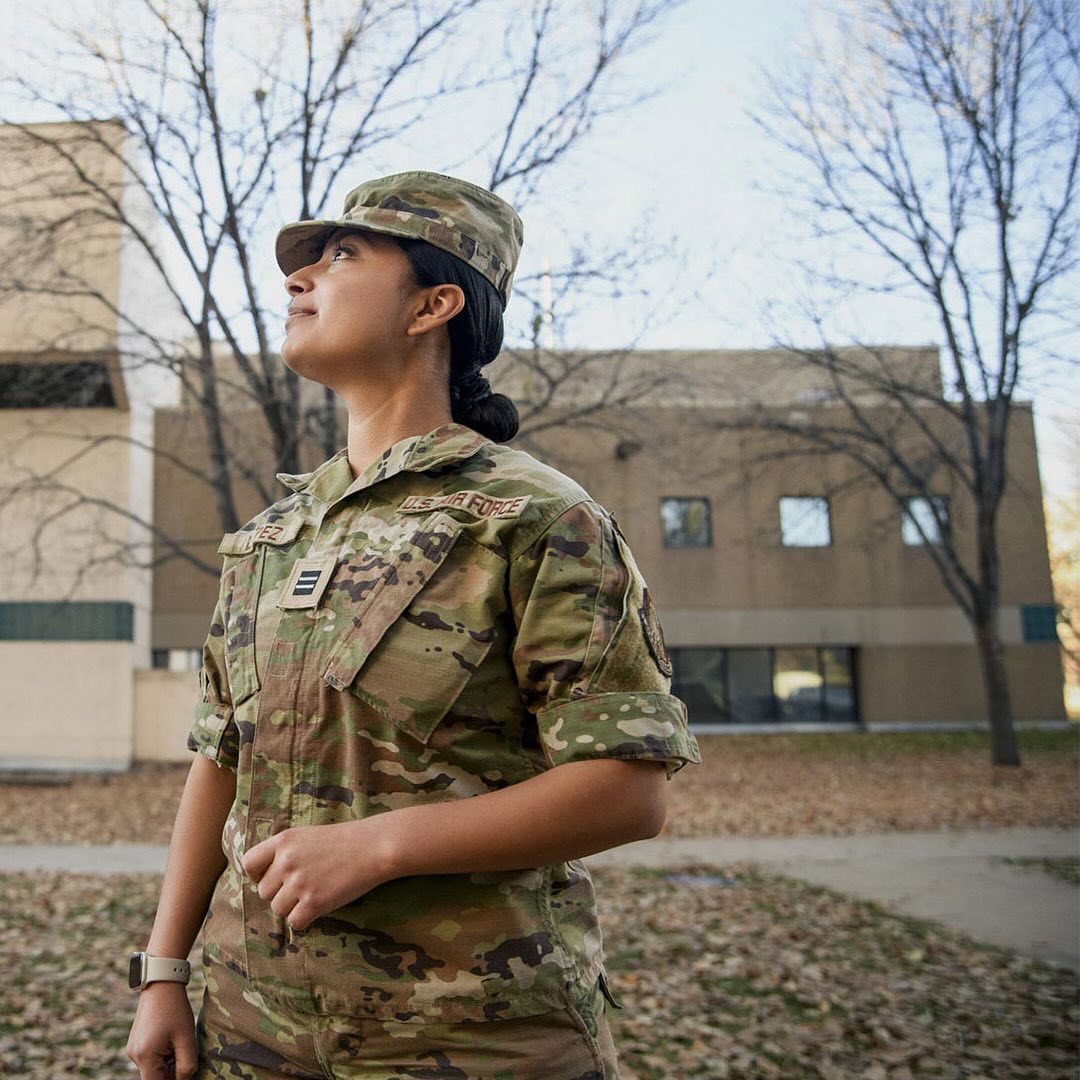 A woman in military uniform, wearing a camouflage cap, stands outside a building under a blue sky with bare trees.