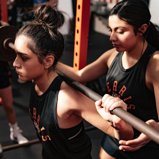 Two women in a gym; one is about to squat with a barbell and the other is spotting. They wear matching tank tops with text.