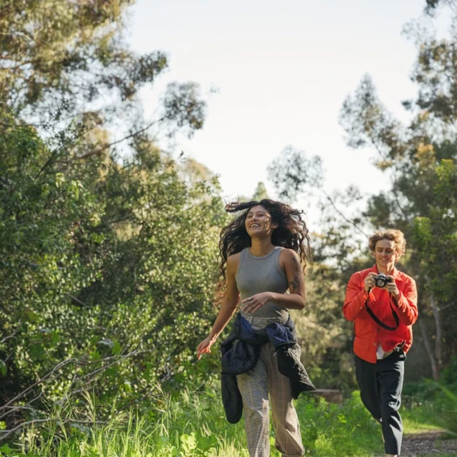 Two women smiling while walking outside through a green, tree-lined path; one takes a picture with a camera, the other has her arms outstretched.