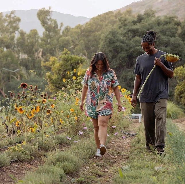 A woman in a floral romper walks with a man carrying a sunflower in a garden setting with trees and mountains in the background.