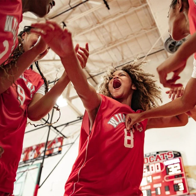 Female basketball team celebrates on the court in red jerseys. Teammates in a huddle with arms raised, showing the team's excitement and unity.