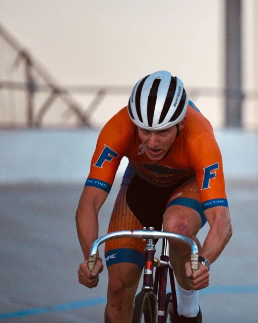 A focused cyclist in orange and blue, wearing a white helmet, powerfully pedals a track bike with silver handlebars on an indoor velodrome.