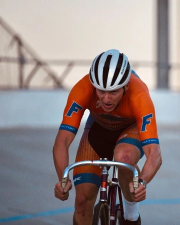 A focused cyclist in orange and blue, wearing a white helmet, powerfully pedals a track bike with silver handlebars on an indoor velodrome.