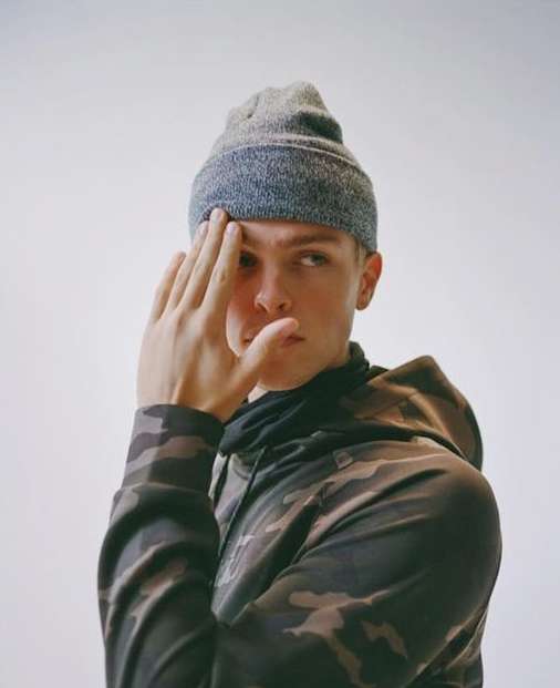 Portrait of a young adult male wearing a grey beanie and camouflage print hoodie, covering part of his face with his hand indoors.