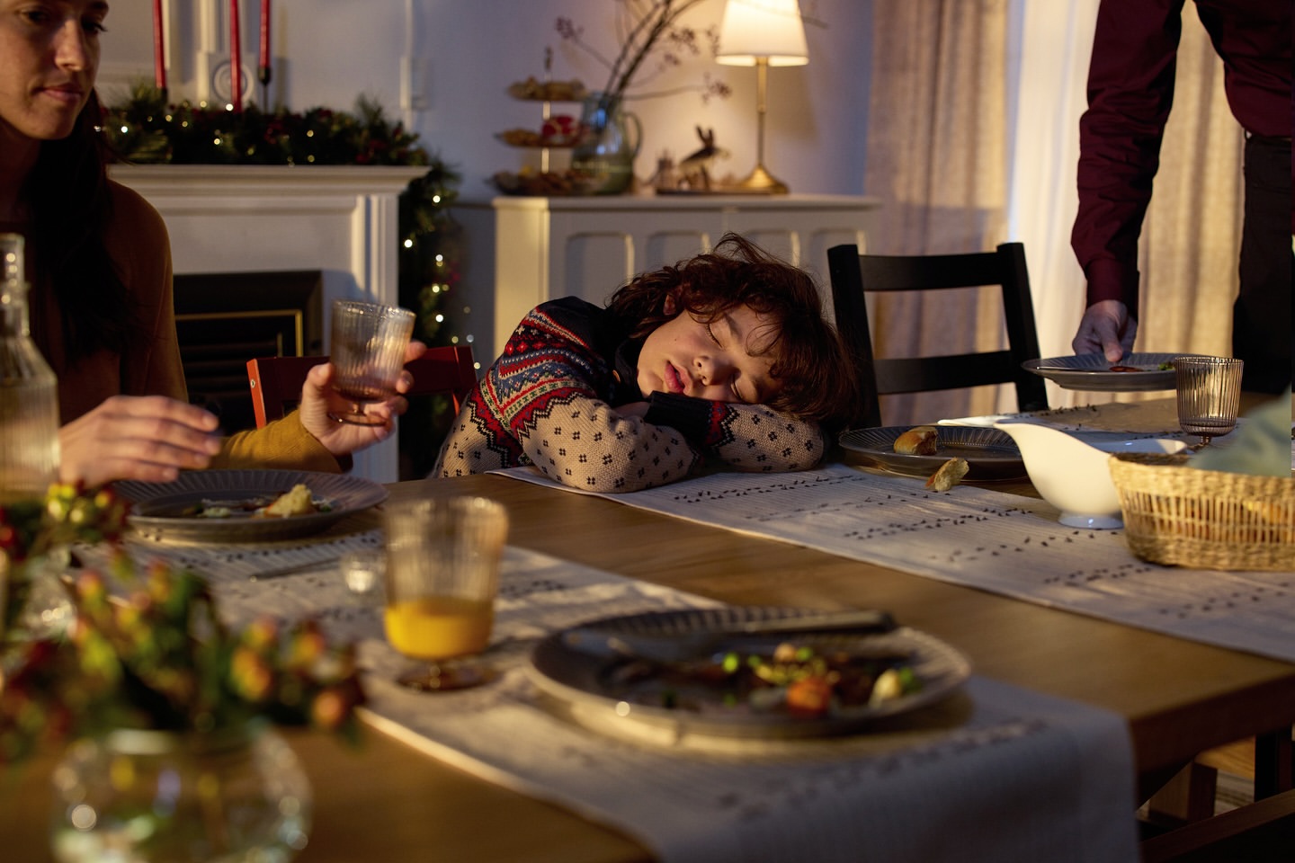 A child sleeps at a table set for a holiday meal as adults mill around. Warm lighting creates a cozy, festive atmosphere in the home.