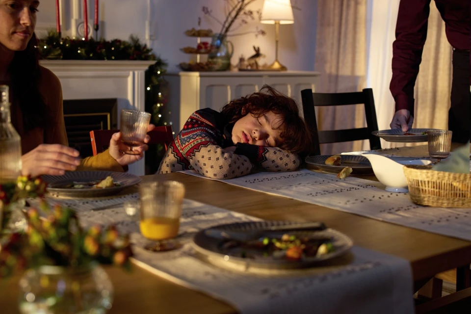 A child sleeps at a table set for a holiday meal as adults mill around. Warm lighting creates a cozy, festive atmosphere in the home.