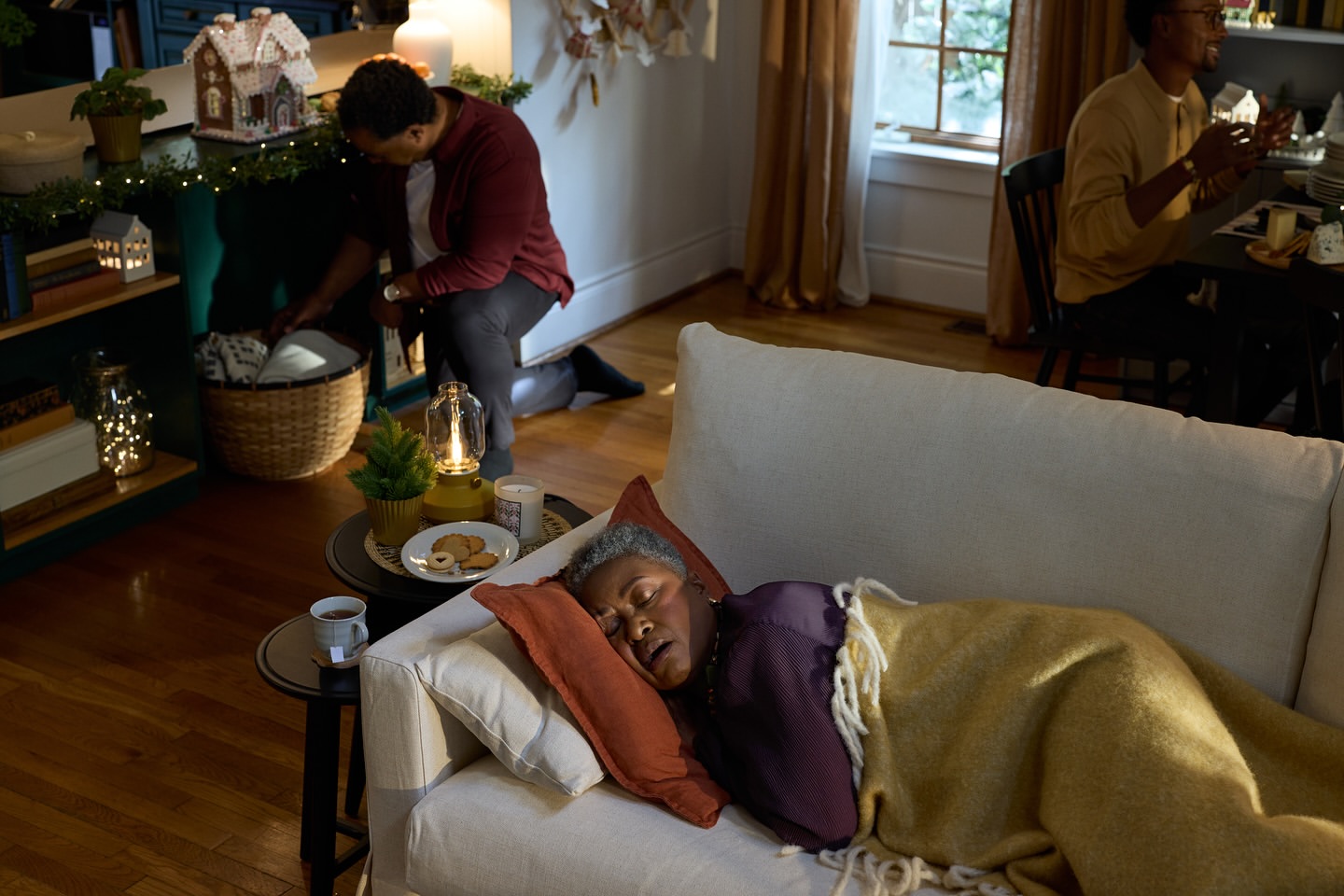 Mature African-American woman napping on couch with blanket and pillow in cozy, warmly-lit interior, with two men in background.