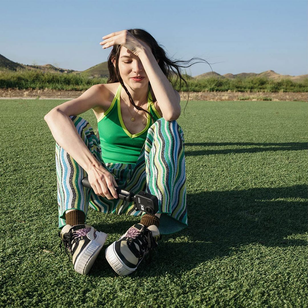 Woman sitting on a turf field shields her eyes from the sun while holding a camera; mountains and blue sky are in the distance.