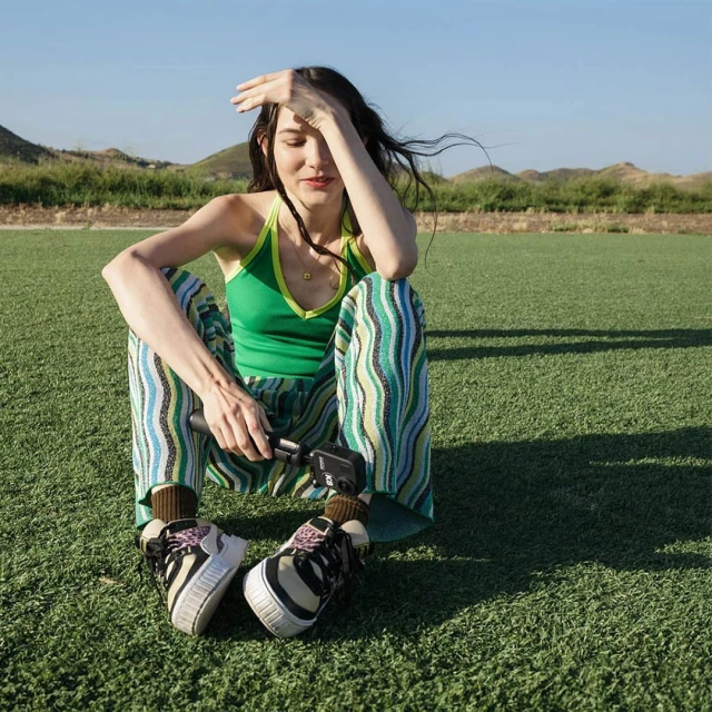 Woman sitting on a turf field shields her eyes from the sun while holding a camera; mountains and blue sky are in the distance.