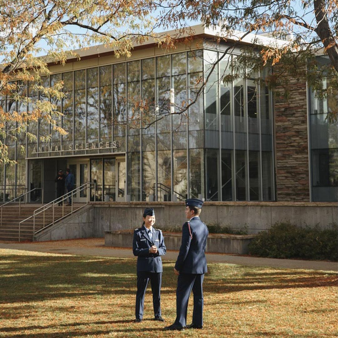 Two individuals in Air Force uniforms stand on a lawn near the 'Animal Sciences' building, surrounded by autumn trees and natural light.