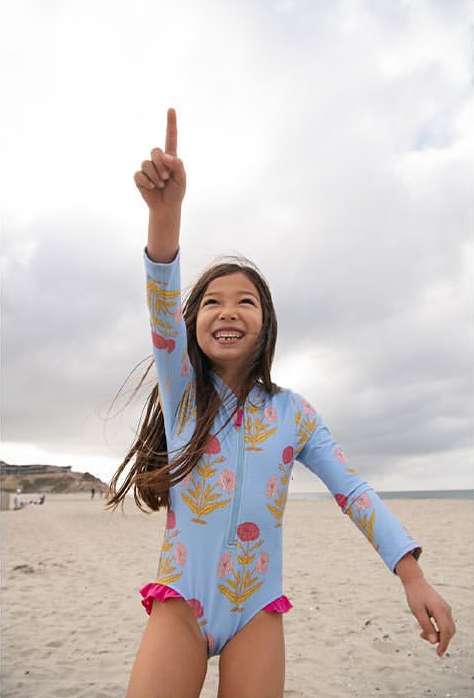 A young girl in a patterned swimsuit stands on a sandy beach under a cloudy sky, pointing upwards with a big smile on her face.