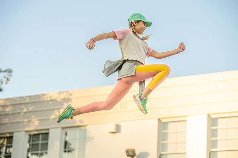 A young girl leaps in the air with a joyful expression, wearing sportswear and a cap, against a backdrop of a building and blue sky.