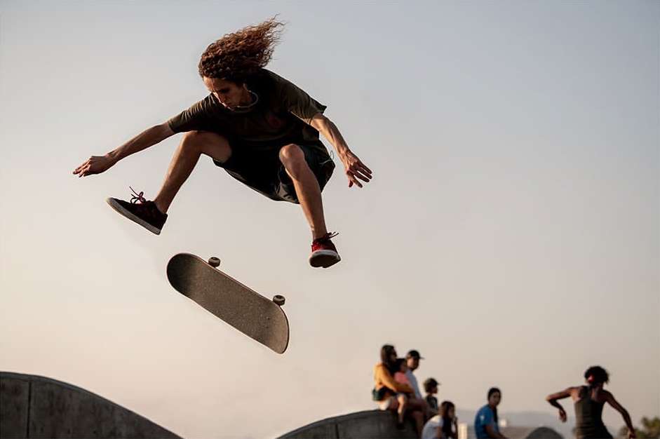 Adult male skateboarder in mid-air performing a trick on a skateboard in front of other people sitting outdoors during the daytime.