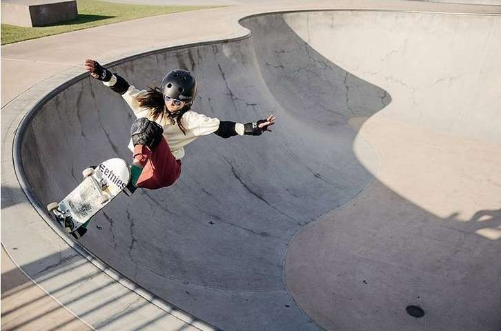 A woman skateboarding in a concrete skatepark bowl, wearing protective gear, a white shirt, and reddish pants.