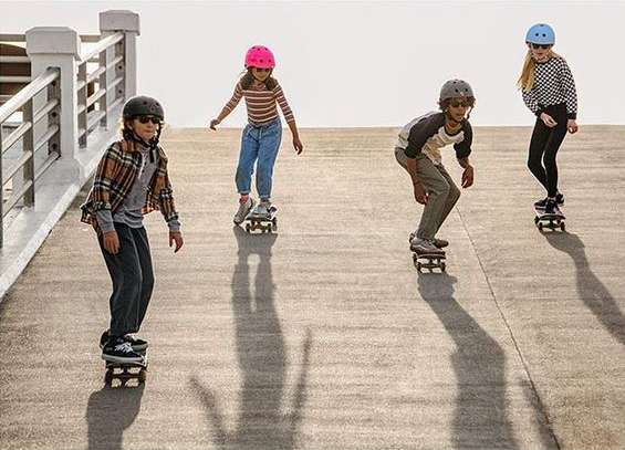 Four children skateboarding outdoors on a sunny day. Kids are wearing helmets and casual clothing, and are on skateboards on a paved surface.