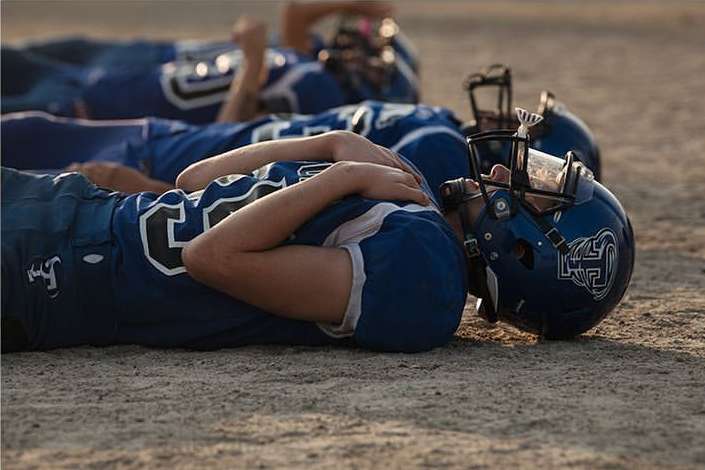 Three American football players in blue uniforms and helmets are lying on a dirt field, resting after practice, with sunlight casting shadows.