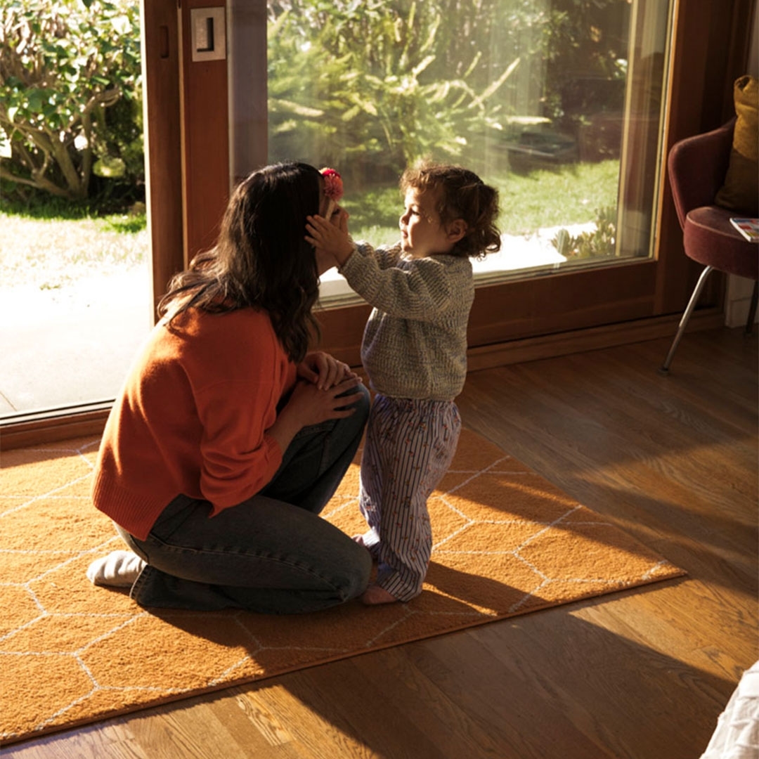 A toddler playfully touches an adult woman's face near a window, creating a heartwarming indoor scene with natural light on a yellow rug.
