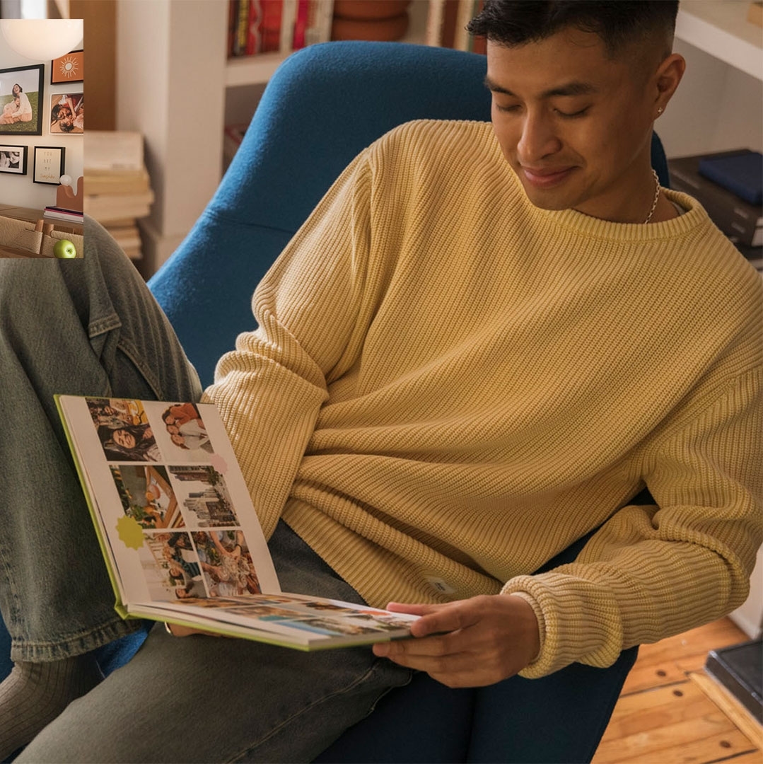 Young man sitting indoors, reading an album with a light yellow ribbed sweater and jeans; bookshelves and framed pictures in the background.