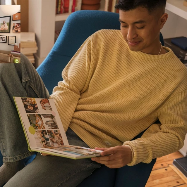 Young man sitting indoors, reading an album with a light yellow ribbed sweater and jeans; bookshelves and framed pictures in the background.