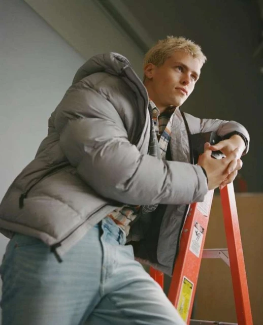 A blonde male model in a gray puffer jacket and light-wash jeans leans on an orange ladder inside a photography studio. 