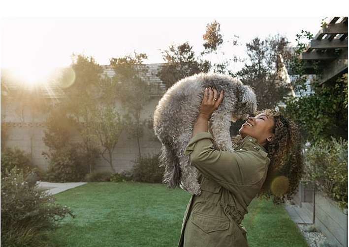 Woman in olive jacket smiles while holding gray poodle outdoors in natural lighting with greenery in the background on a sunny day.