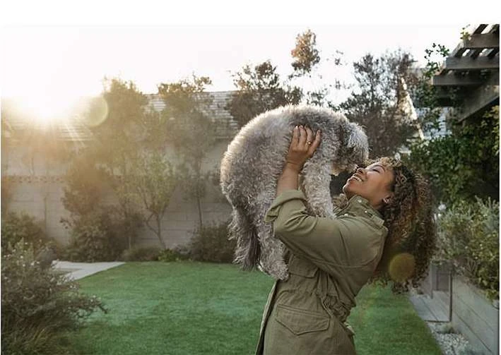 Woman in olive jacket smiles while holding gray poodle outdoors in natural lighting with greenery in the background on a sunny day.
