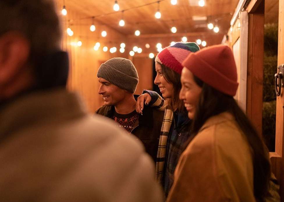 Three young adults in colorful beanies smile at an unseen person at an outdoor gathering, under warm string lights and wood walls.