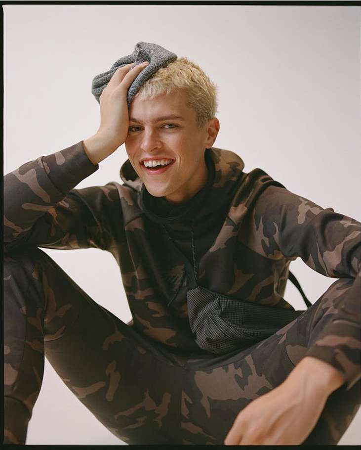 Blond male model in camouflage sweat suit smiles in a studio, holding a gray towel on his head, set against a neutral backdrop.