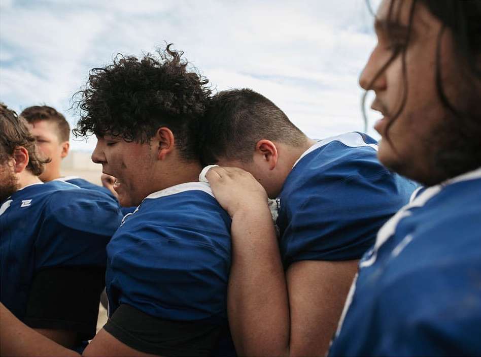 Close-up of football players embracing in blue and white uniforms. Intense emotions, dirt on faces. Cloudy outdoor setting, teamwork.