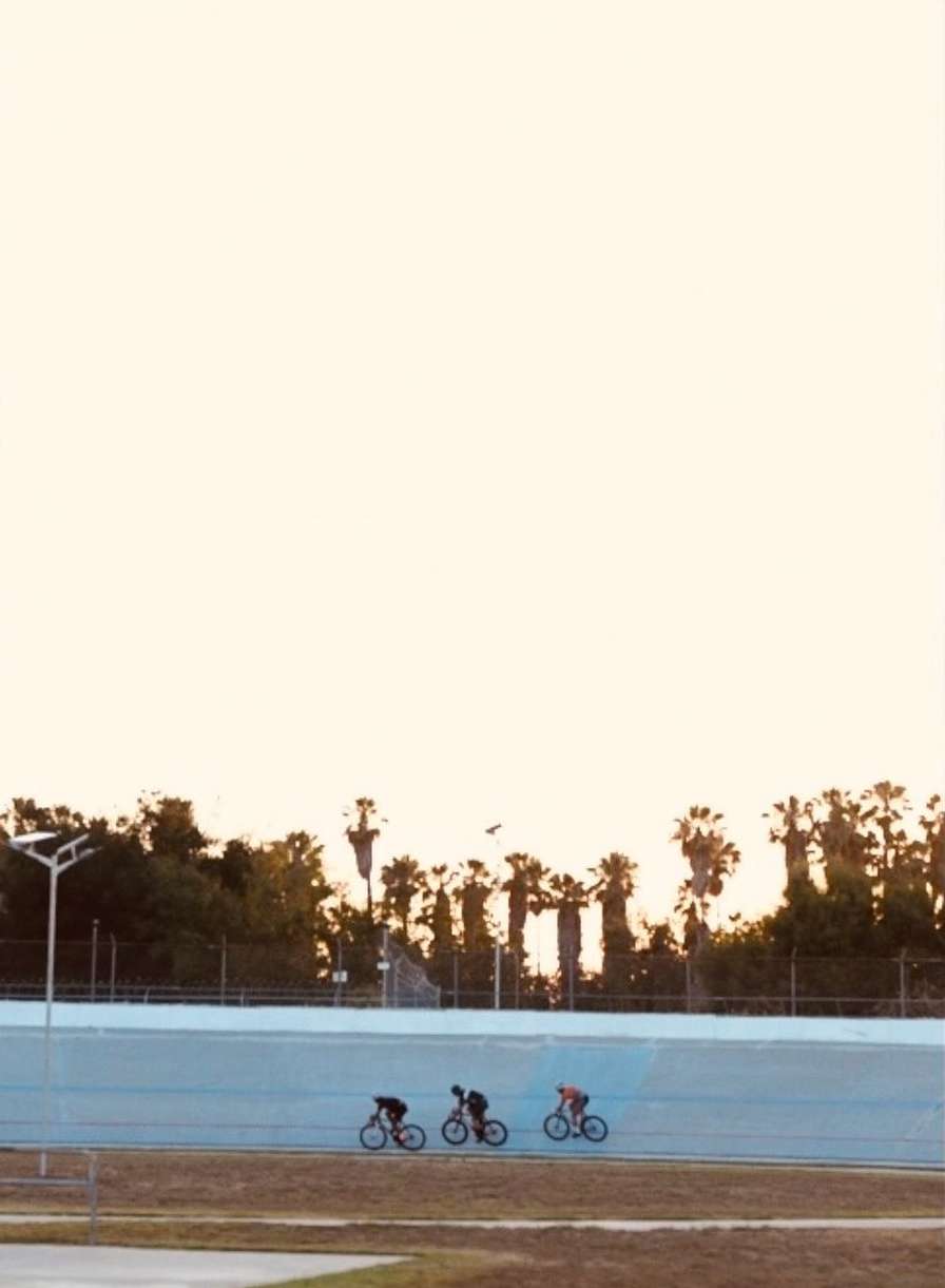 Three cyclists ride bikes on a track near palm trees at sunset. Cyclists wear dark clothes; the track is blue and brown with an open sky.