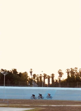 Three cyclists ride bikes on a track near palm trees at sunset. Cyclists wear dark clothes; the track is blue and brown with an open sky.