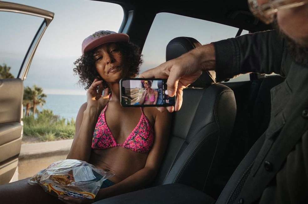 Woman in bikini poses in car while being photographed with a phone against ocean background. Summer vacation travel lifestyle.