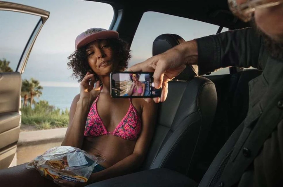 Woman in bikini poses in car while being photographed with a phone against ocean background. Summer vacation travel lifestyle.