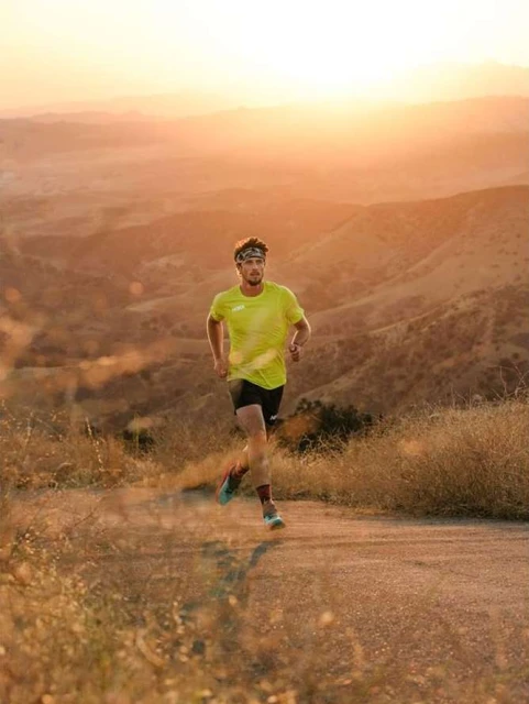 A man in sportswear runs on a dirt path, silhouetted by a golden sunset over hills in the background. He is focused and athletic.