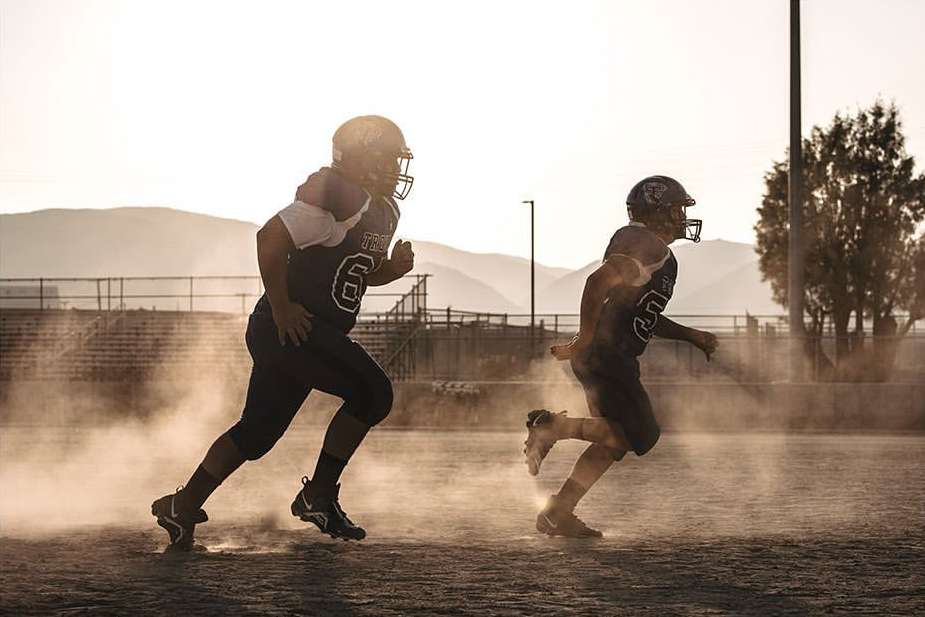 Two American football players in uniform running on a field with a backdrop of mountains, illuminated by sunlight and dust.