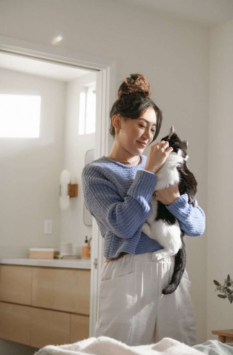 Woman with a bun hairstyle smiles while holding a black-and-white cat inside an airy room with soft, natural lighting.