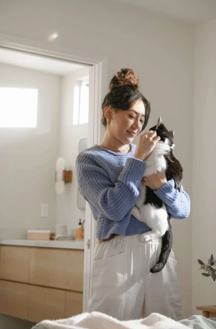 Woman with a bun hairstyle smiles while holding a black-and-white cat inside an airy room with soft, natural lighting.