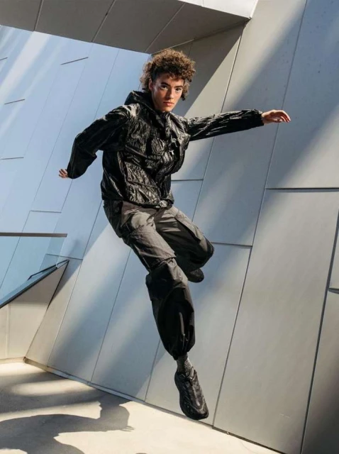 Athletic young adult in black hooded jacket and cargo pants jumping against an architectural backdrop with shadows and highlights on concrete.
