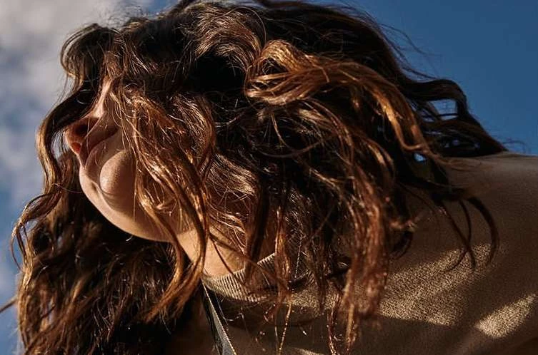 Close-up shot of a brunette adult female looking upwards against a bright blue sky, warm lighting accentuating wavy hair texture.