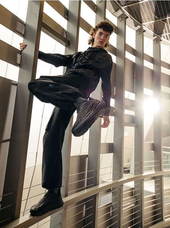 Stylish man in dark clothing poses confidently on a metal railing in a sunlit, modern architectural space, showcasing fashion and attitude.