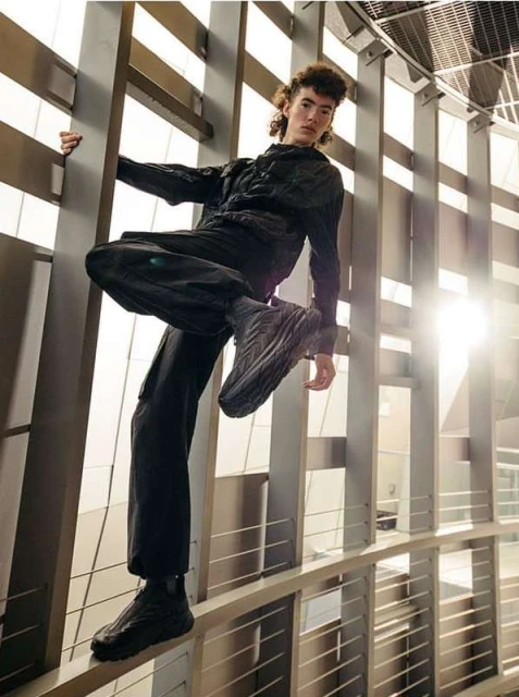 Stylish man in dark clothing poses confidently on a metal railing in a sunlit, modern architectural space, showcasing fashion and attitude.